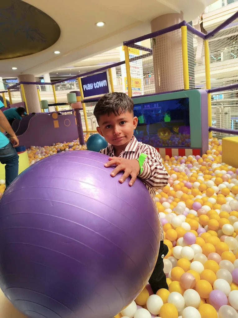 A young child holds a large purple ball in a ball pit at a play center.