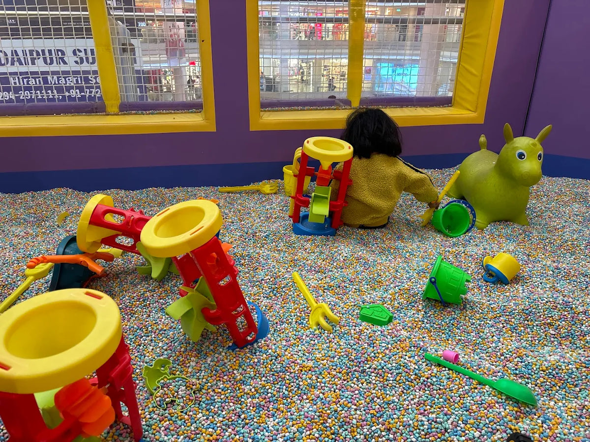 A young child plays in a ball pit with colorful toys, including a green ride-on toy.