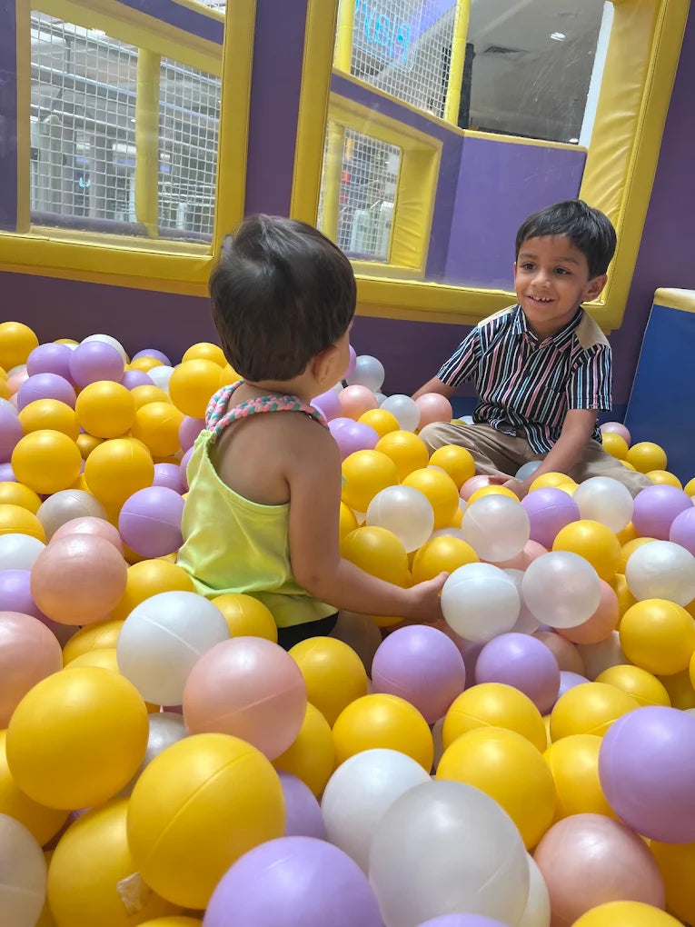 Two young children play in a ball pit filled with yellow, purple, and white balls.