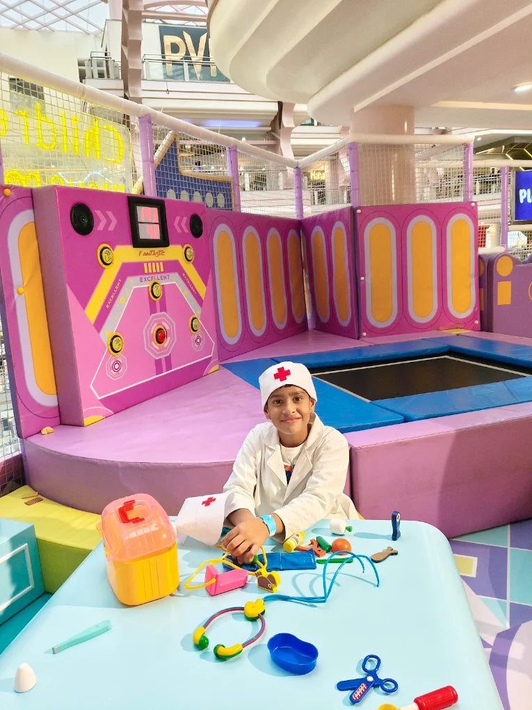 A young child dressed as a doctor plays with toy medical equipment in a colorful indoor play area.