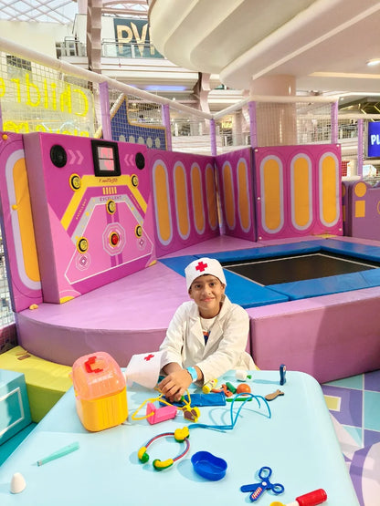 A young child dressed as a doctor plays with toy medical equipment in a colorful indoor play area.