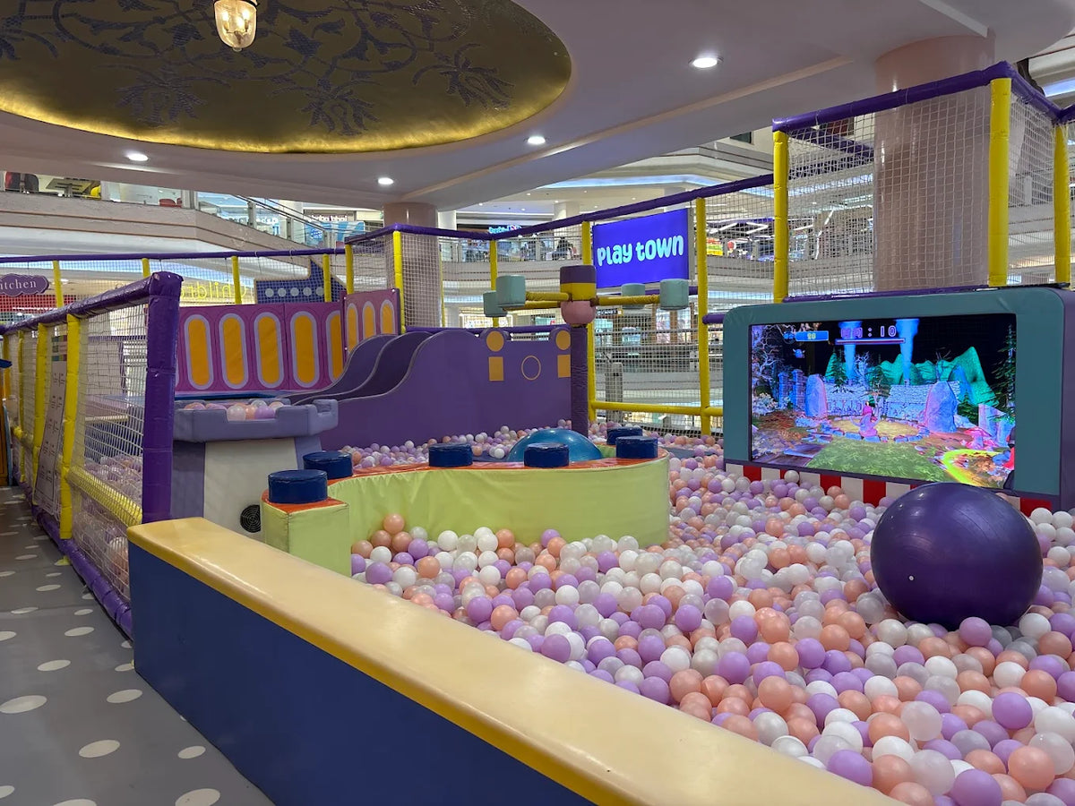 A colorful indoor playground with a ball pit and slides, featuring a "Play Town" sign and a screen displaying a game.
