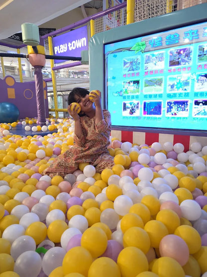 A young child plays in a ball pit in front of a large screen displaying games.