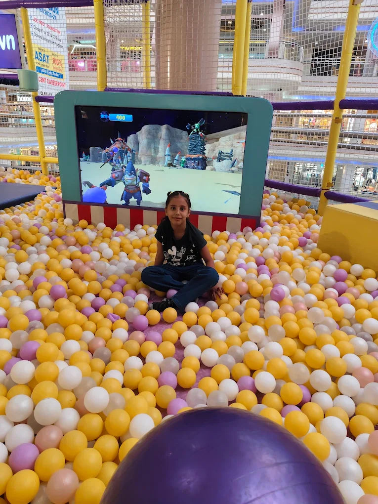 A young girl sits in a ball pit in front of a video game screen.