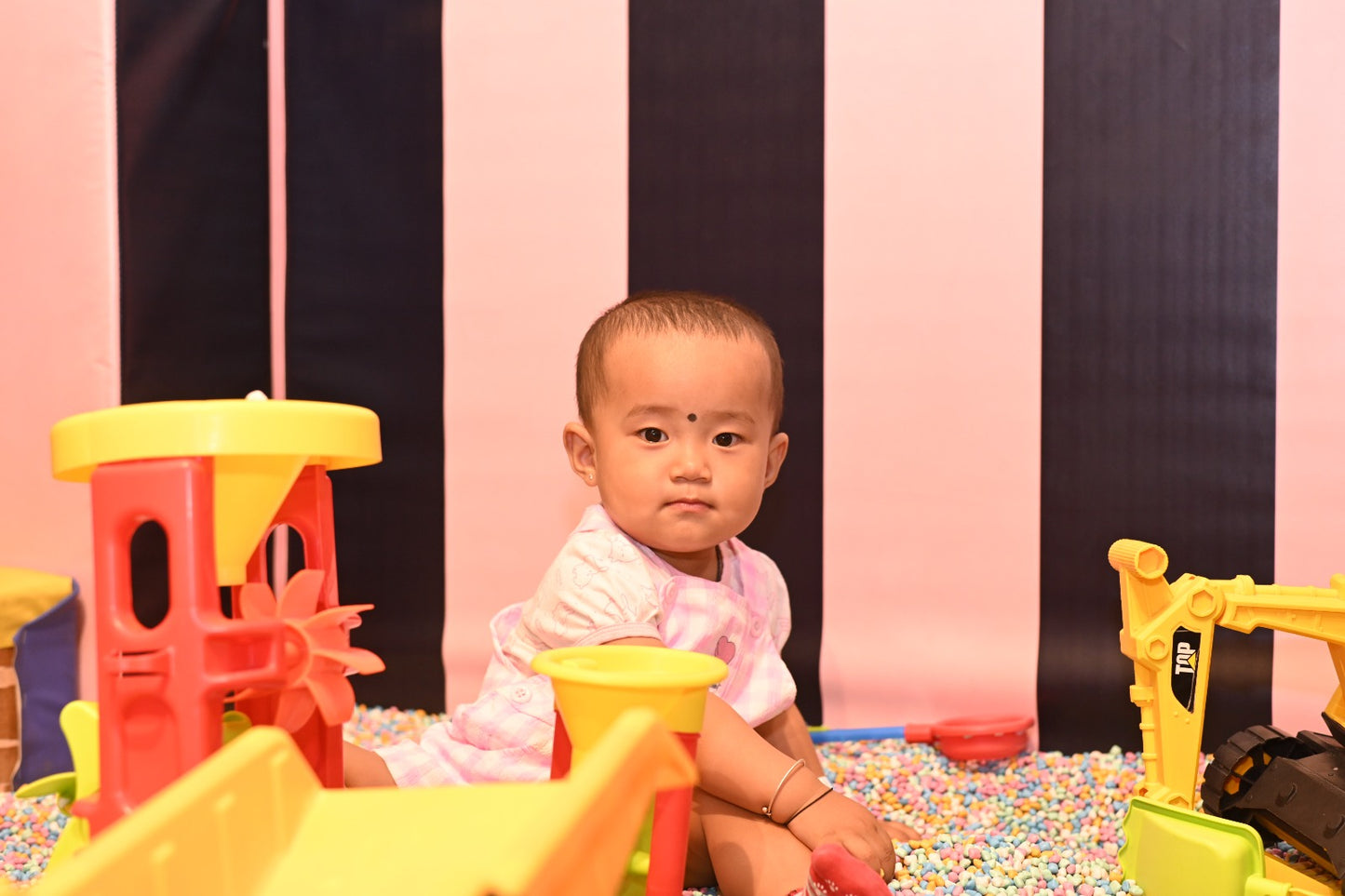 A baby sits in a ball pit surrounded by colorful toys, with a striped backdrop.