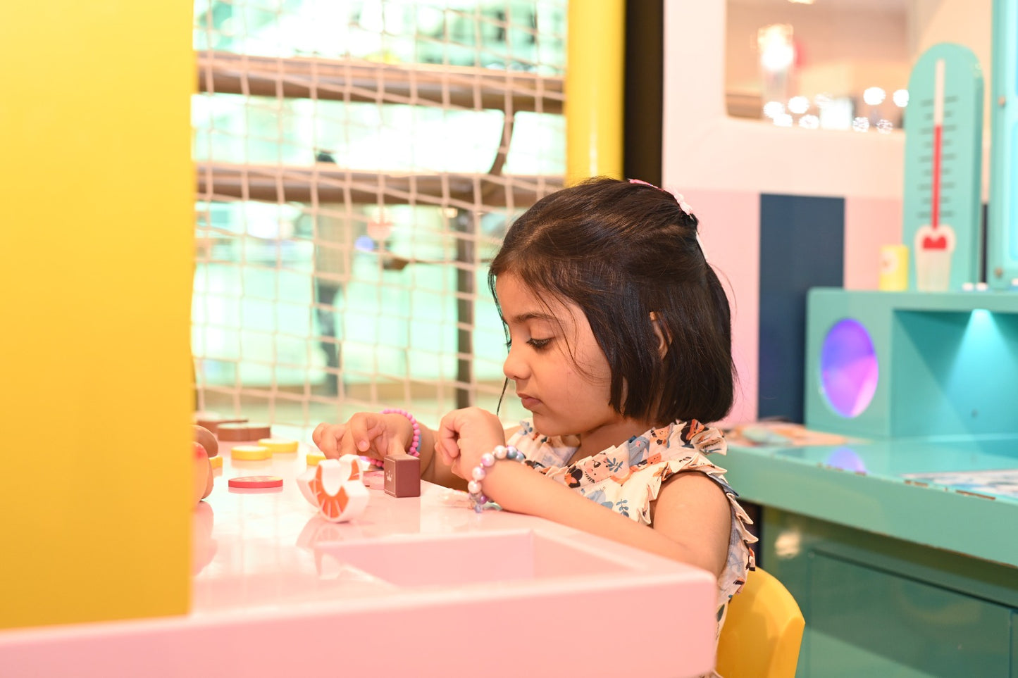 A young girl plays with colorful blocks at a pink table in a bright, playful indoor setting.