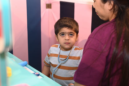 A young boy wearing a toy stethoscope looks up as a woman in a purple shirt stands nearby.