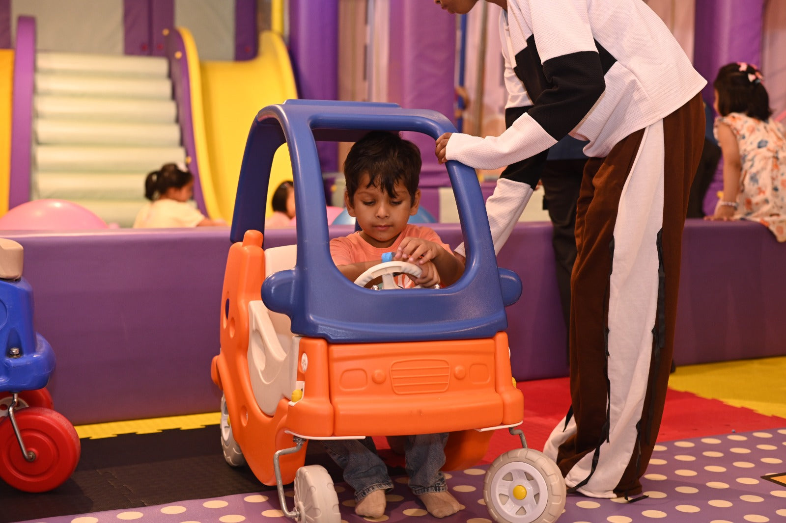 A young boy plays in a toy car at an indoor playground while an adult stands nearby.