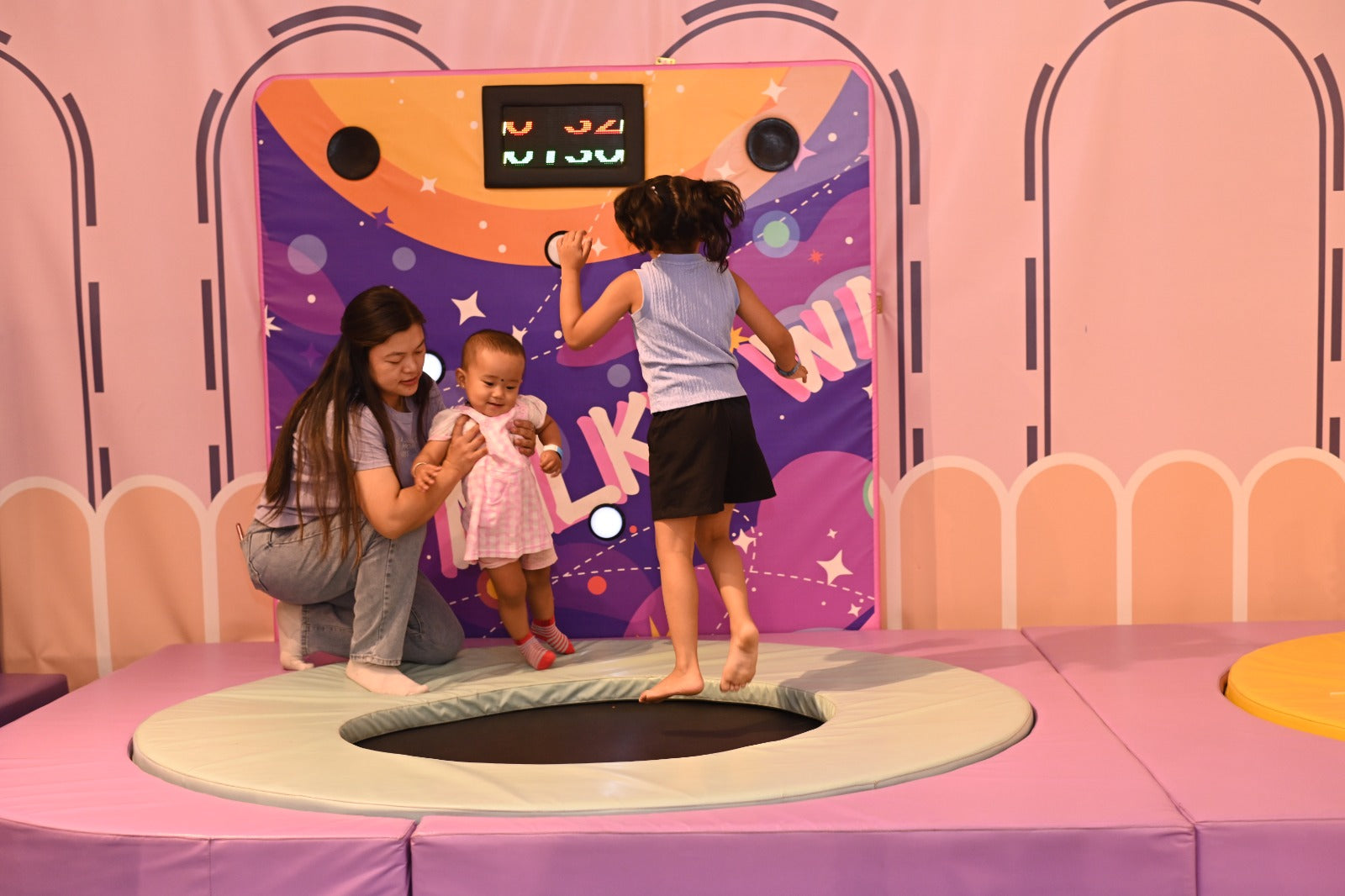 A woman helps a baby on a trampoline while a young girl jumps.