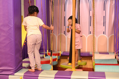 Two young girls navigate a colorful indoor obstacle course with ropes and padded platforms.