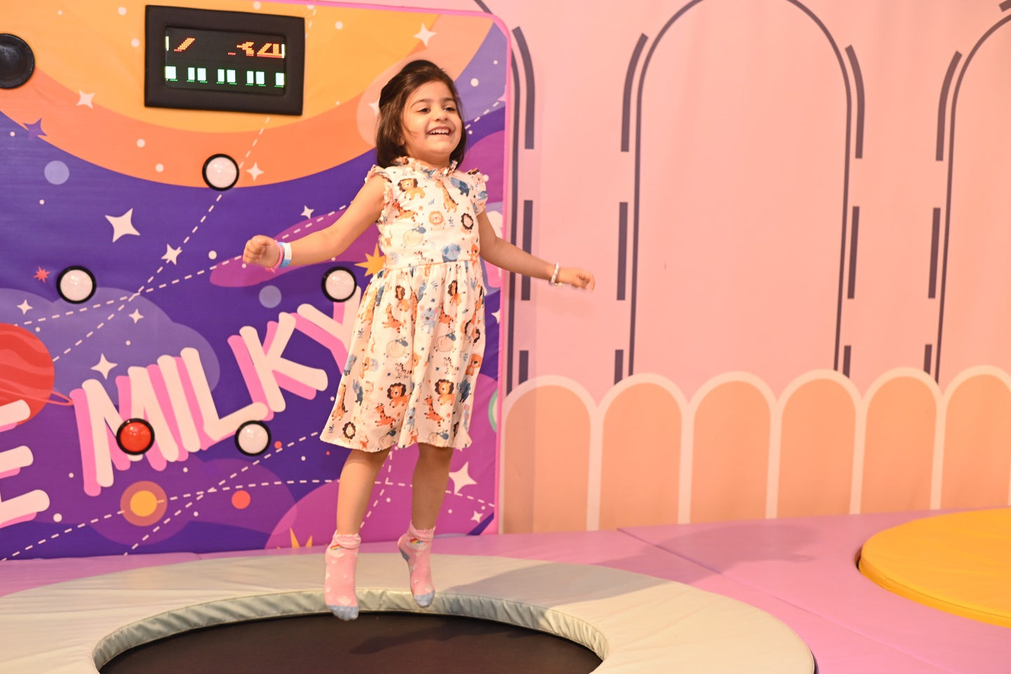A young girl jumps on a trampoline in a colorful indoor play area. She wears a patterned dress and pink socks.