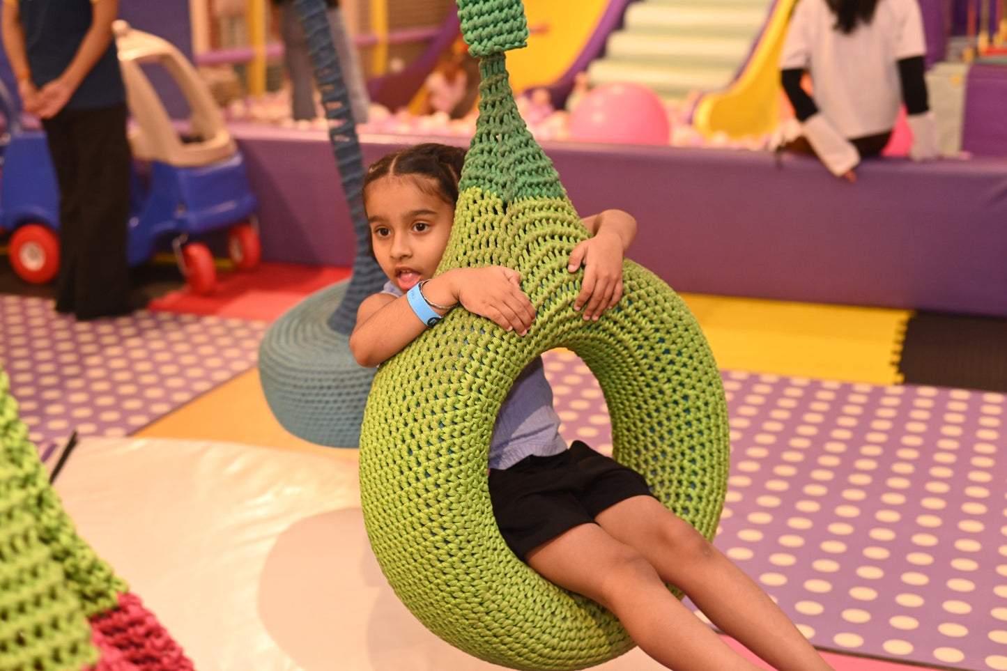 A young girl sits in a green, woven tire swing in a colorful indoor play area.