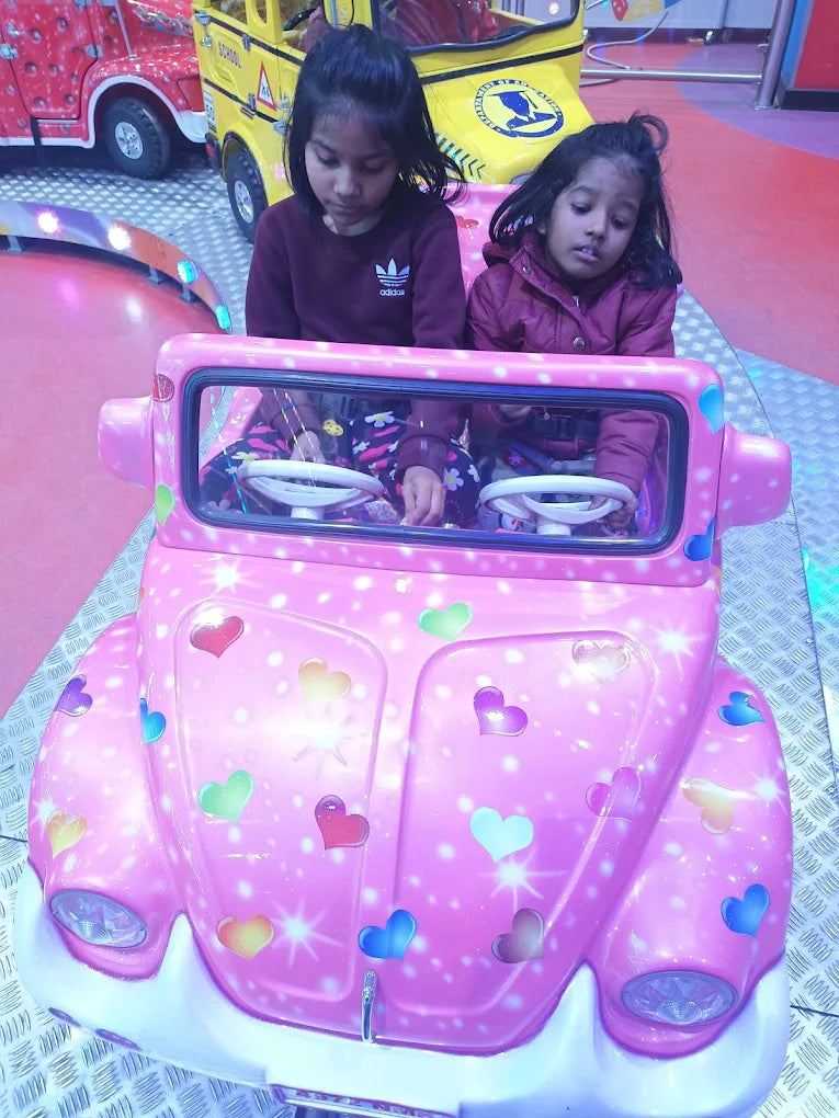 Two young girls ride in a pink toy car at an amusement park.