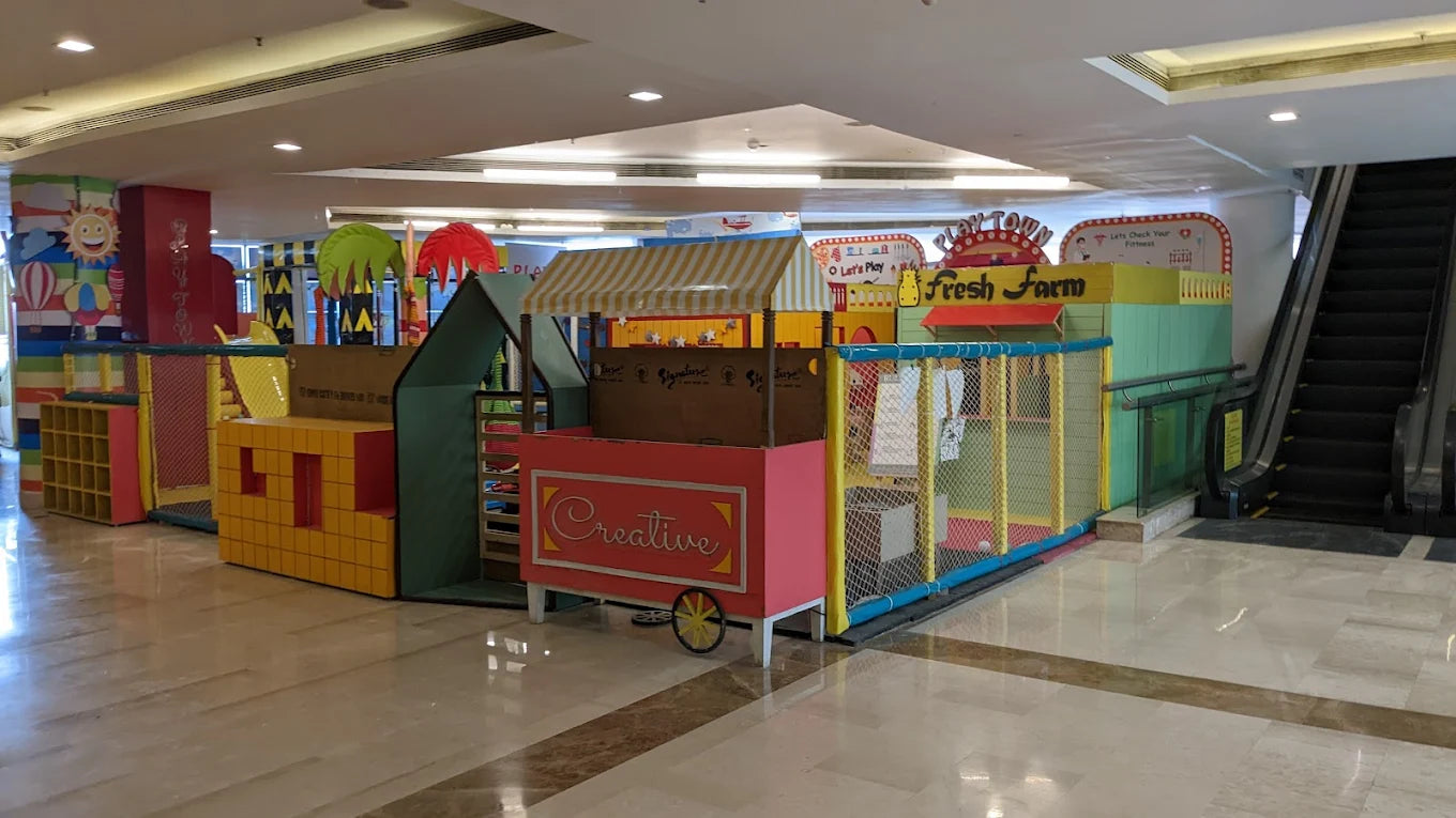 A colorful indoor playground with a "Fresh Farm" theme, featuring soft play structures and a small cart.