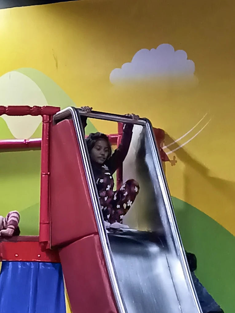 A young girl sits at the top of a red and silver slide, ready to go down. The background is a brightly colored wall with cartoon clouds.