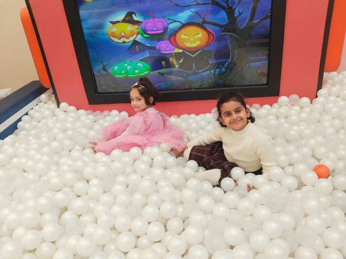 Two young girls play in a ball pit in front of a Halloween-themed screen.