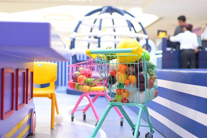 Children's play shopping carts filled with toy produce are in a bright, indoor play area.