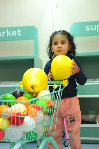 A young child holds a large yellow ball while standing next to a toy shopping cart filled with colorful plastic fruits.