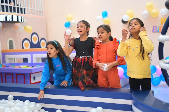 Four young girls in colorful outfits play with white balls in a ball pit at a party.