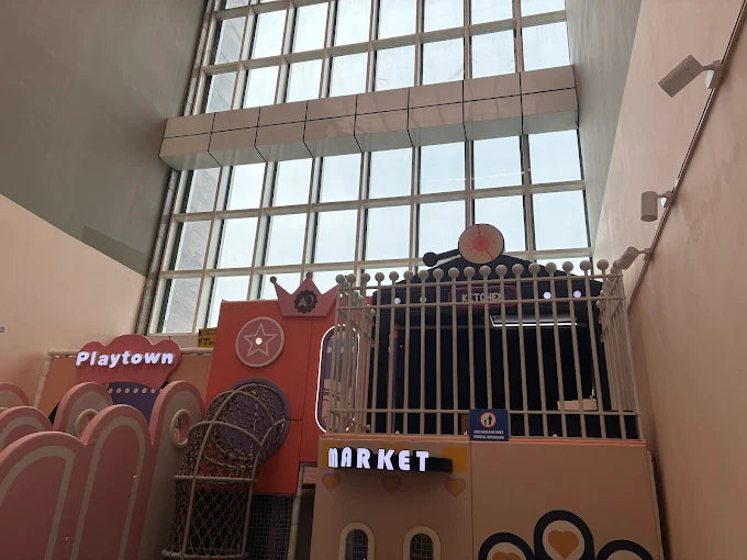 A colorful indoor playground with "Playtown" and "Market" signs, featuring a kitchen area and a large window.