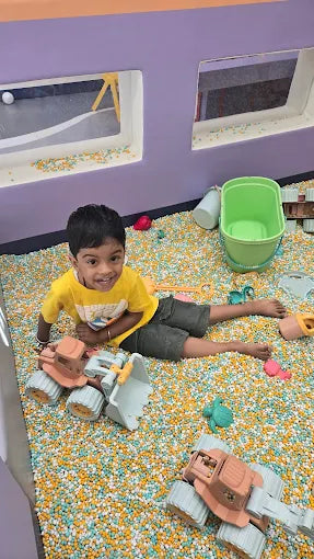 A young boy plays with toy trucks in a ball pit filled with colorful beads.