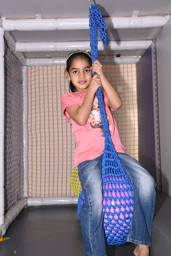 A young girl in a pink shirt and jeans swings on a blue net swing in an indoor play area.