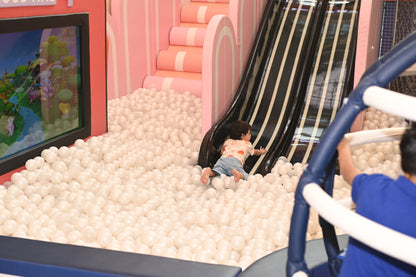A toddler plays in a ball pit next to a slide, with another child in the foreground.