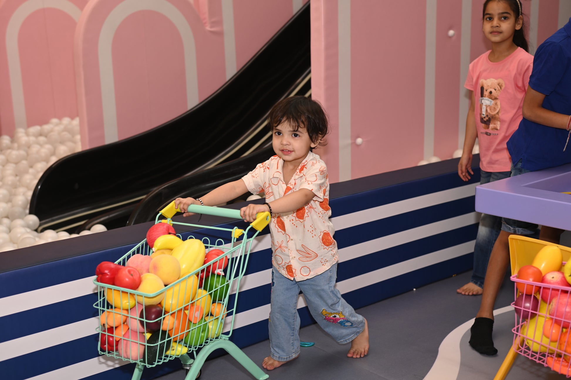 A young child pushes a toy shopping cart filled with plastic fruit in a play area with a ball pit and slide.