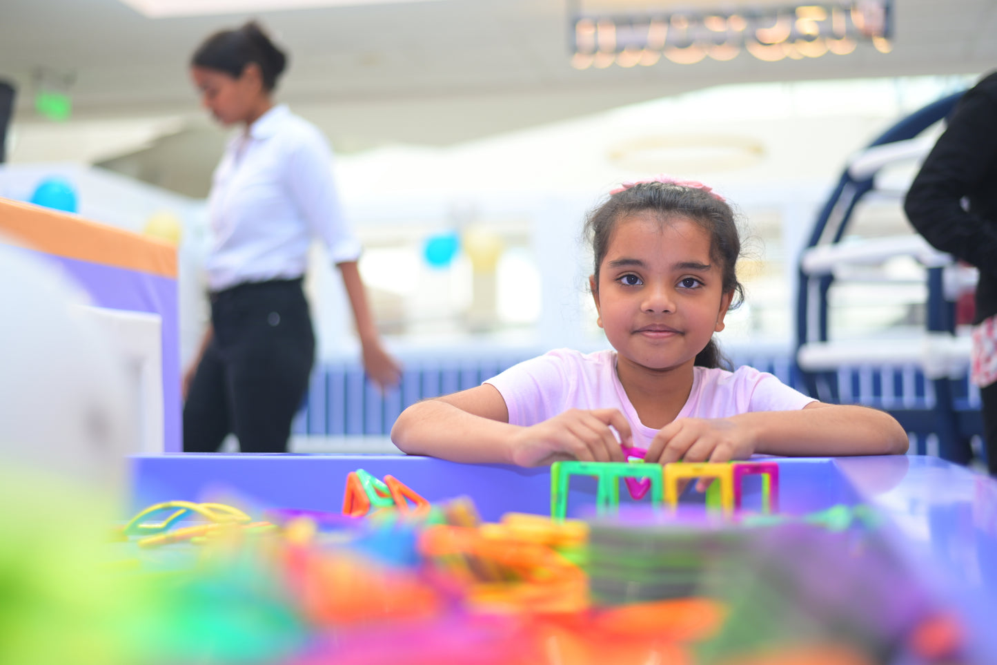 A young girl plays with colorful toys at a table, with another person blurred in the background.