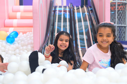 Two young girls smile while playing in a ball pit with a colorful slide in the background.