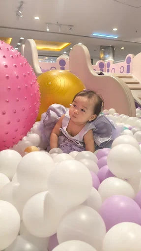 A baby sits in a ball pit with large pink and yellow balls in the background.