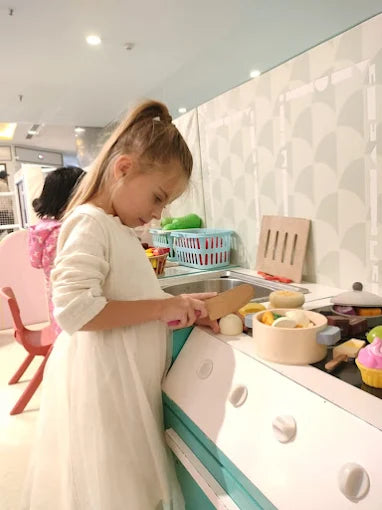 A young girl in a white dress plays with a toy kitchen, chopping food on a cutting board.