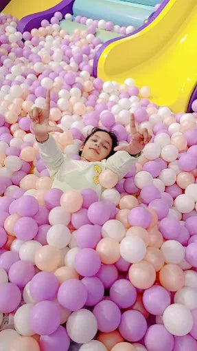A young child lies in a ball pit, surrounded by pastel-colored balls, with a yellow slide in the background.