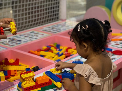 A young child plays with colorful building blocks at a table. Another child's hands are visible, holding a yellow gear.