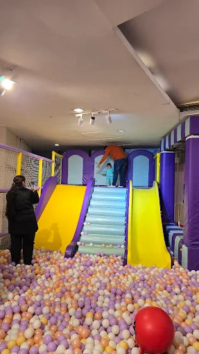 A child plays in a ball pit with adults nearby, near slides and stairs in an indoor playground.