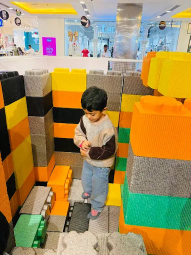 A young child stands in a play area made of large, colorful building blocks. The child is wearing a sweater and jeans.