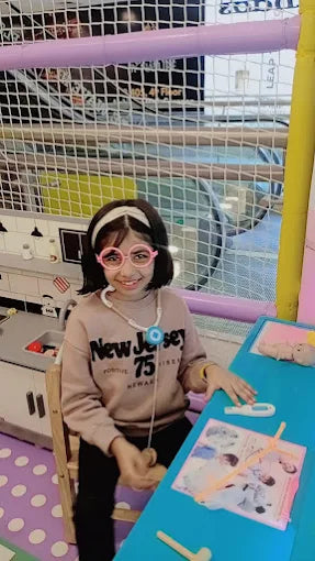 A young girl in pink glasses plays doctor at a blue table in a play area.