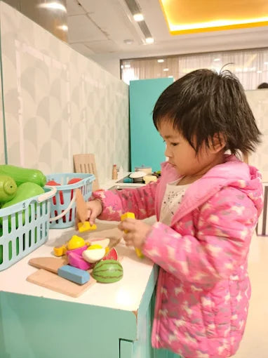 A young child in a pink jacket plays with toy fruits and vegetables on a counter.