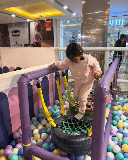 A young child plays in a ball pit, balancing on a tire swing.