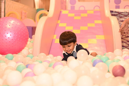 A young boy smiles while playing in a ball pit with a pink slide in the background.