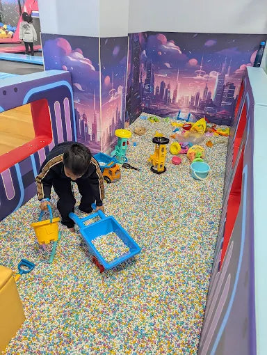 A young child plays in a ball pit with a blue wagon and yellow bucket.