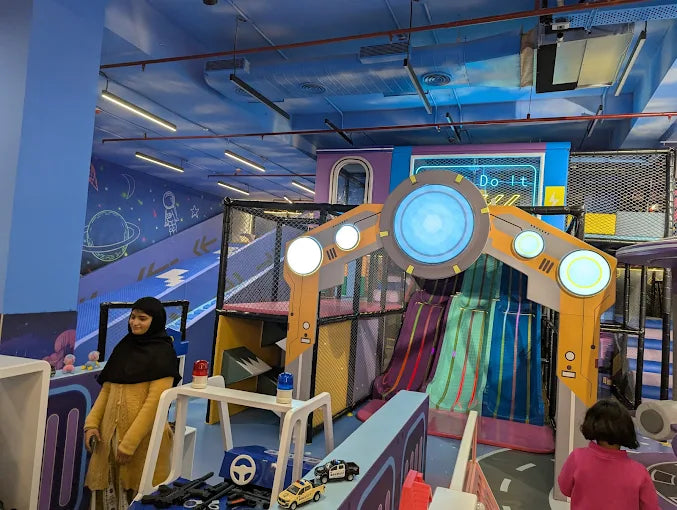 A young girl in a yellow cardigan and headscarf stands in a colorful indoor playground with slides and a space-themed mural.