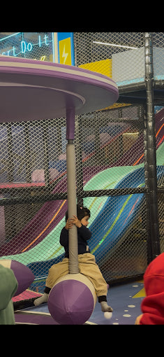 A child sits on a pole in a colorful indoor playground with slides in the background.