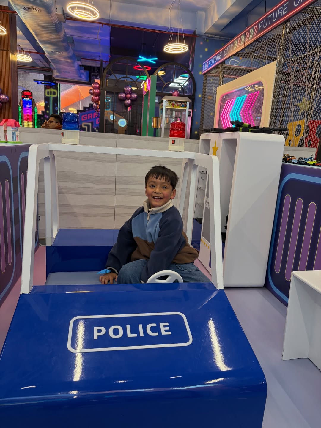 A young boy smiles while sitting in a toy police car at an indoor amusement park.