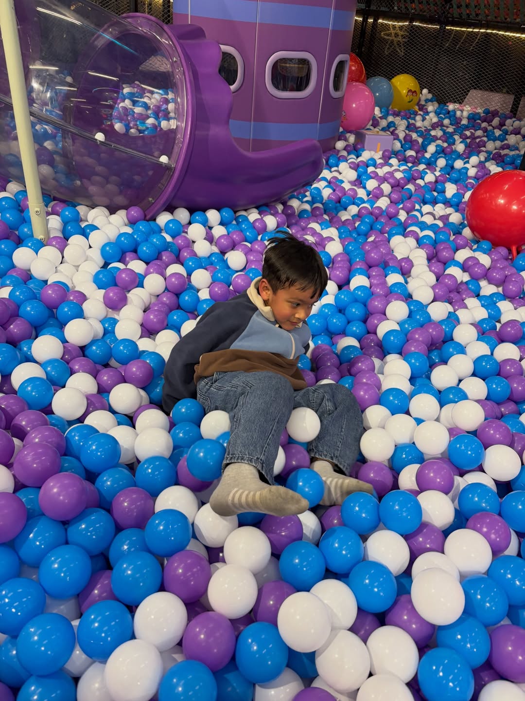 A young boy sits in a ball pit filled with purple, blue, and white balls, with a purple play structure behind him.