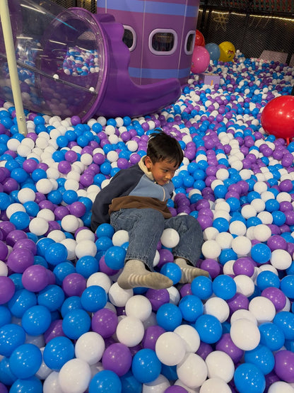 A young boy sits in a ball pit filled with purple, blue, and white balls, with a purple play structure behind him.