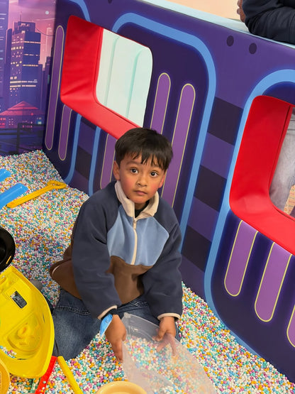 A young boy sits in a ball pit, playing with a clear container of colorful balls. He is in a playful, indoor setting with a train-themed backdrop.