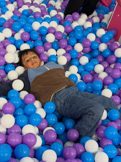 A young child lies in a ball pit filled with blue, purple, and white balls.