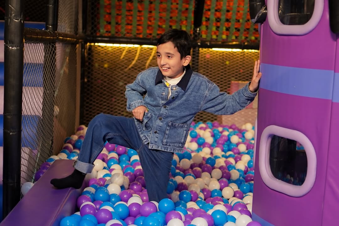 A young boy in a denim jacket and jeans plays in a ball pit. He is leaning against a purple structure.