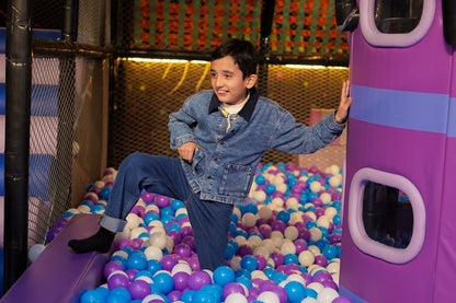 A young boy in a denim jacket and jeans plays in a ball pit. He is leaning against a purple structure.